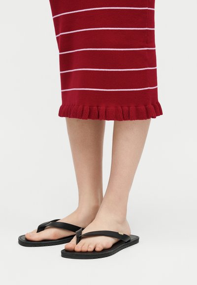 Πόδια που φορούν κόκκινη φούστα με λευκές ρίγες και βολάν στο τελείωμα, μαύρα επίπεδα σανδάλια τύπου φλιπ-φλοπ σε λευκό φόντο.