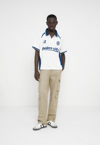 Λευκό πόλο πουκάμισο με μπλε λεπτομέρειες και το κείμενο "palm city", σε συνδυασμό με μπεζ παντελόνι cargo και μαύρα-άσπρα αθλητικά παπούτσια.