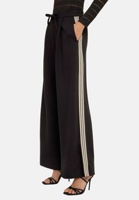 Svart byxor med breda ben och dragsko i midjan, med vertikala vita ränder på sidorna och sidofickor. Slät tygtextur.