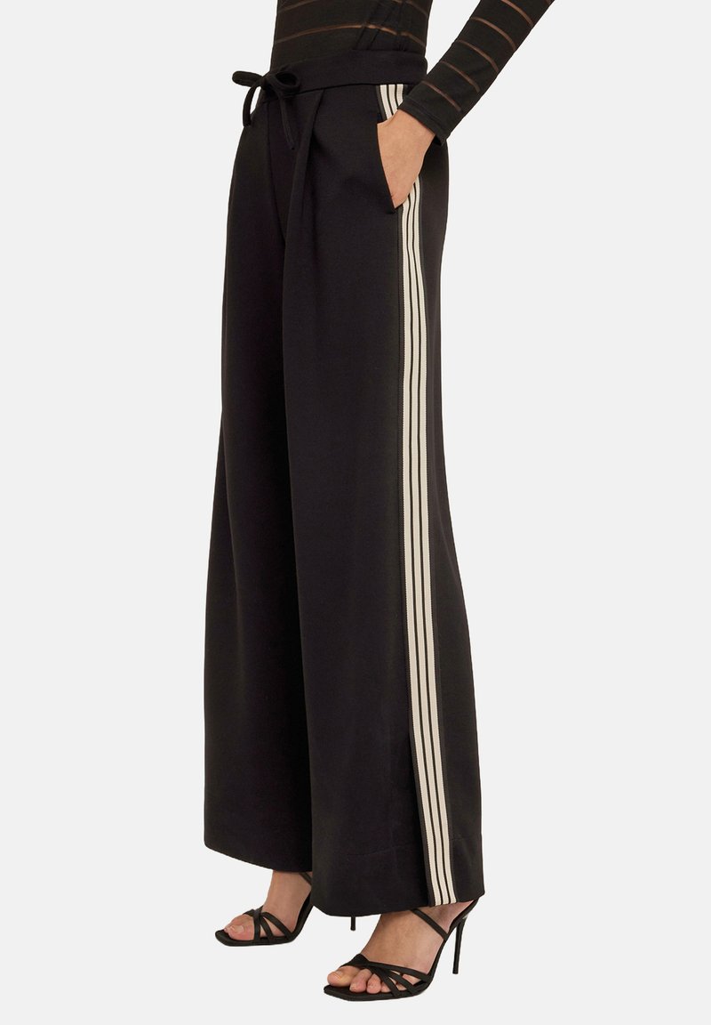 Svart byxor med breda ben och dragsko i midjan, med vertikala vita ränder på sidorna och sidofickor. Slät tygtextur.