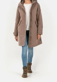 Parka larga beige con capucha, frontal con cremallera, bolsillos laterales, cordones ajustables, llevada sobre una blusa blanca y vaqueros azules, combinada con botas marrones.