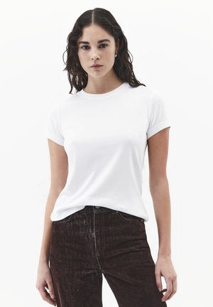 Junge Frau mit langen dunklen Haaren, die ein schlichtes weißes Kurzarm-T-Shirt trägt, das in hoch taillierte dunkelbraune Hosen gesteckt ist.