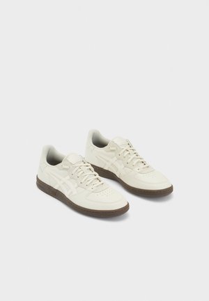 Paire de baskets en cuir blanc cassé avec des rayures beige clair et des semelles en caoutchouc marron, présentées côte à côte sur un fond uni.