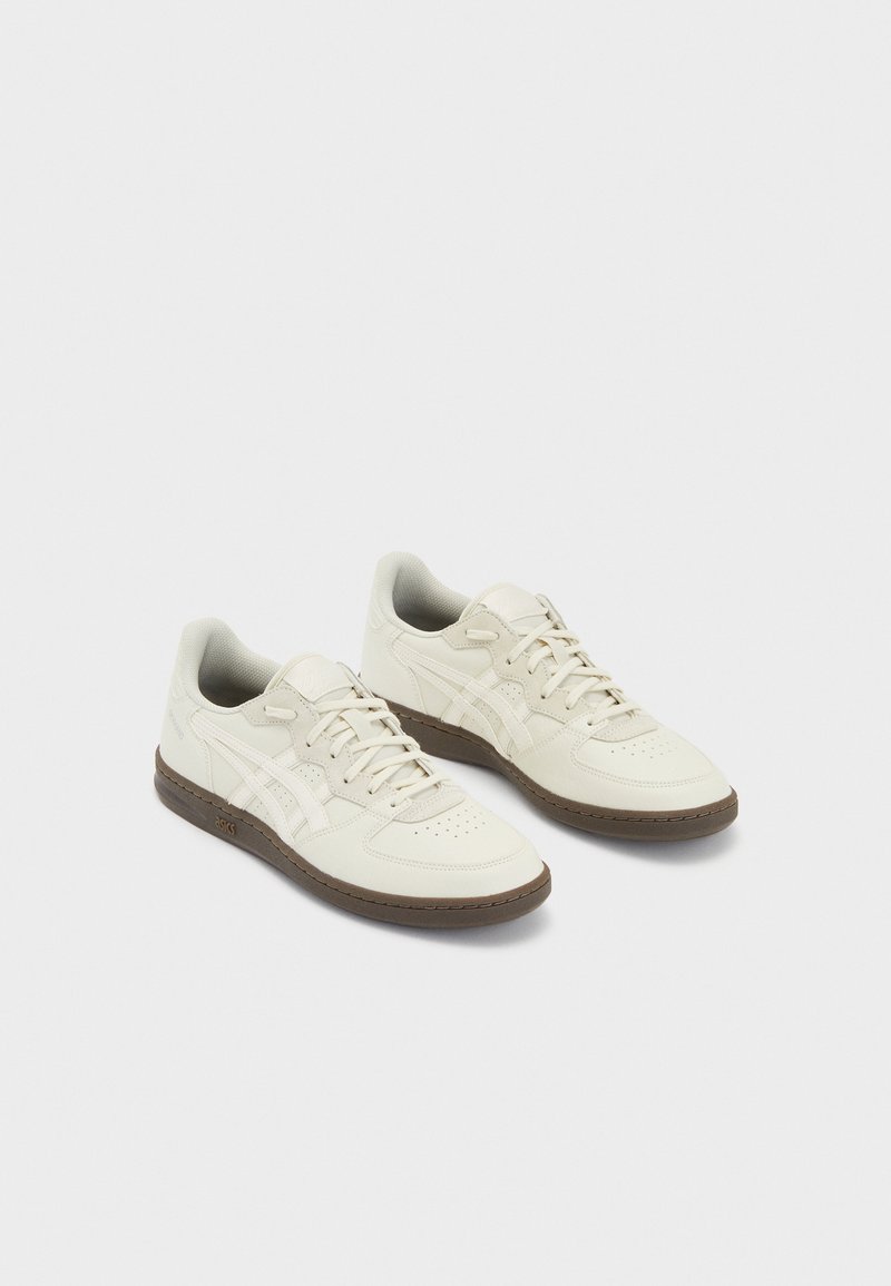 Paire de baskets en cuir blanc cassé avec des rayures beige clair et des semelles en caoutchouc marron, présentées côte à côte sur un fond uni.