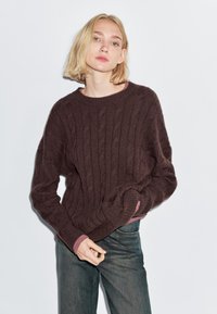 Maglione marrone a maglia cable con collo rotondo e maniche lunghe; presenta dettagli rosa ai polsini. Indossato con jeans in denim scuro.