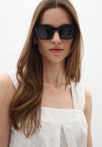 Lunettes de soleil noires angulaires avec des montures épaisses. Le modèle porte un haut blanc texturé avec des bretelles. L'arrière-plan est neutre et minimaliste.