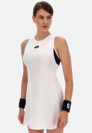 Vrouw draagt een mouwloze witte tennisjurk met zwarte zijkanten en zwarte polsbandjes, kijkt opzij tegen een effen achtergrond.