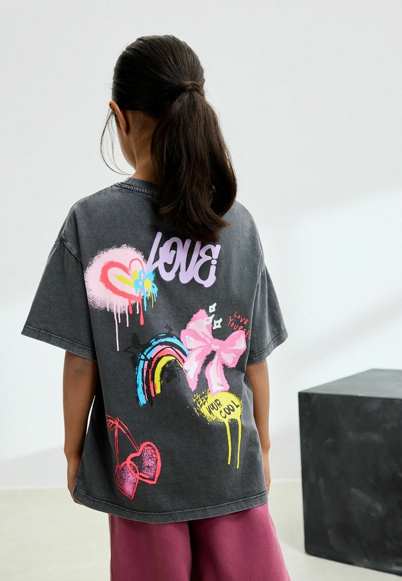 Ragazza con capelli scuri raccolti in una coda di cavallo che indossa una maglietta grigia con grafica colorata a tema cuore, fiocco, arcobaleno e amore sulla schiena.