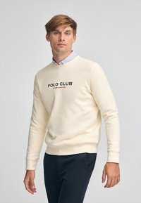 Sudadera crema con puños y dobladillo acanalados, que presenta el logotipo "POLO CLUB" y "ESTABLECIDO 2012" en negro. Usada sobre una camisa de cuello a rayas.