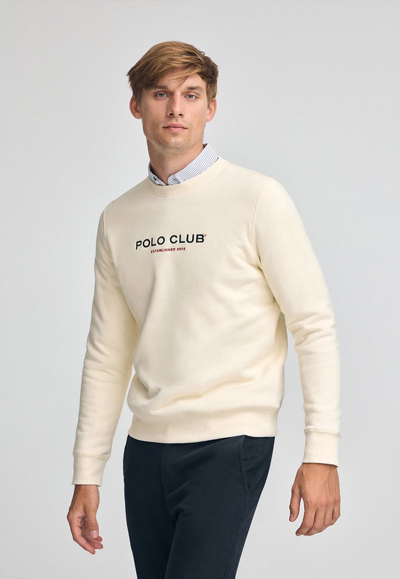 Sudadera crema con puños y dobladillo acanalados, que presenta el logotipo "POLO CLUB" y "ESTABLECIDO 2012" en negro. Usada sobre una camisa de cuello a rayas.