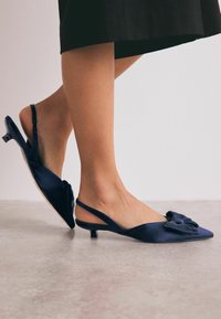 Tummansininen slingback-kengät, joissa on terävä kärki, satiinimateriaali ja koristeellinen nauha etuosassa. Matala kitten-korko.