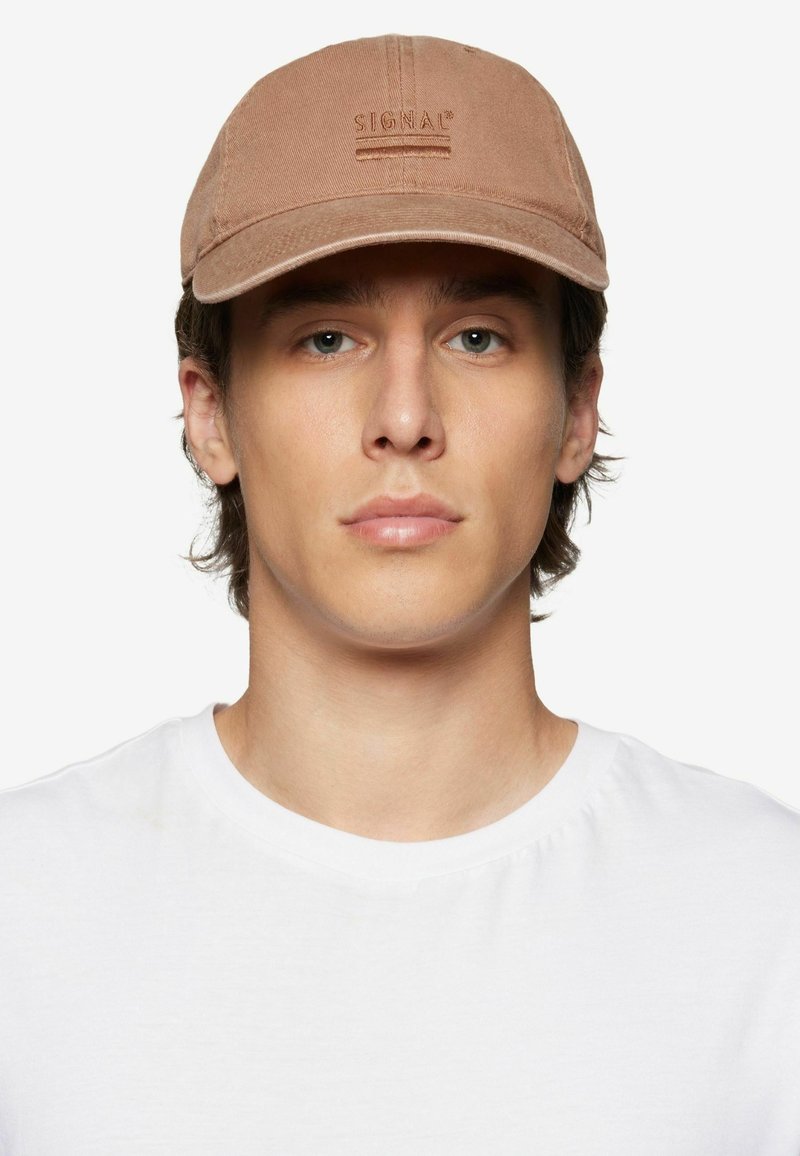 Căciulă din material brown cu visieră curbă și un logo embossat, purtată de o persoană îmbrăcată într-un tricou alb simplu. Vedere frontală, fundal minim.