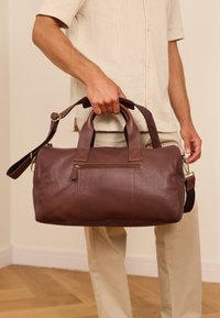 Ruskea nahkainen matkalaukku, jossa on kaksi yläkahvaa, säädettävä olkahihna ja vetoketjullinen tasku. Varustettu teksturoitulla pinnalla ja kullansävyisillä yksityiskohdilla.