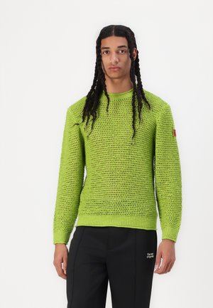 Bright green knit sweater with a loose, open pattern and ribbed cuffs and hem. Worn with black trousers featuring white text detail.