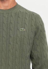 Grønn kabelstrikket genser med rund hals og liten brodert grønn krokodille-logo på venstre bryst.