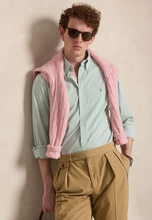 Ung mand iført solbriller, grønstribet skjorte, lyserød sweater hængt over skuldrene og højtaljede beige plisserede bukser.