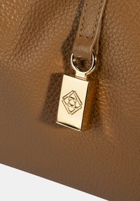 Bruine leren handtas met textuur, voorzien van een gouden charme met een geometrisch design en een lus voor bevestiging. Detaillering van een soepel leren hengsel zichtbaar.