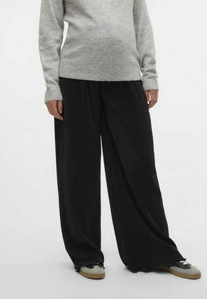 Schwarze, gerippte, weit geschnittene Hosen mit lockerem Sitz, kombiniert mit einem hellgrauen Strickpullover. Die Schuhe haben eine Mischung aus grauen und schwarzen Akzenten.