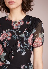 Femme aux cheveux bouclés portant une robe noire à fleurs rouges et bleues avec des détails à volants sur la poitrine et des manches bouffantes transparentes.
