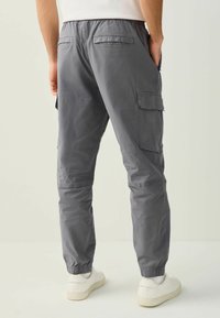 Homme portant un pantalon cargo gris avec des poignets élastiques et des baskets blanches, vu de dos sur un fond uni.