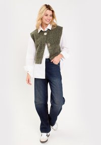 Veste sans manches en laine verte avec de gros boutons, portée par-dessus une chemise blanche, associée à un jean bleu foncé et des baskets blanches.