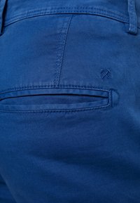 Modré bavlněné kalhoty s zadní kapsou na zip a jemnými vyšitými detaily. Hladká textura a přizpůsobený tvar.