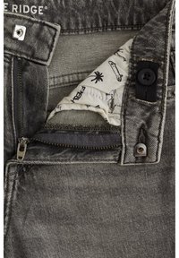 Jean en denim gris avec une fermeture éclair ouverte, doublure en tissu blanc imprimé partiellement visible, bouton noir et fermeture à pression métallique.