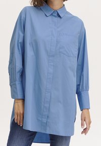 Camicia azzurro chiaro con bottoni, realizzata in cotone, dotata di taschino sul petto, maniche lunghe e vestibilità leggermente oversize; presenta una pattina frontale e maniche con polsini.