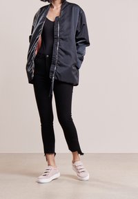 Chaqueta bomber negra de satén con cremallera, llevada abierta sobre una camiseta negra, combinada con jeans negros con dobladillos deshilachados y zapatillas de lona rosa claro.