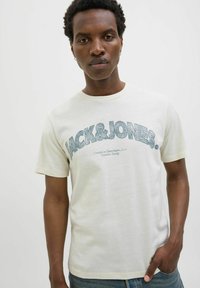 Kremskrāsas kokvilnas T-kreklis ar īsām piedurknēm, kas rotāts ar lielu zilu grafisku attēlu "JACK & JONES" un mazāku tekstu zem tā.