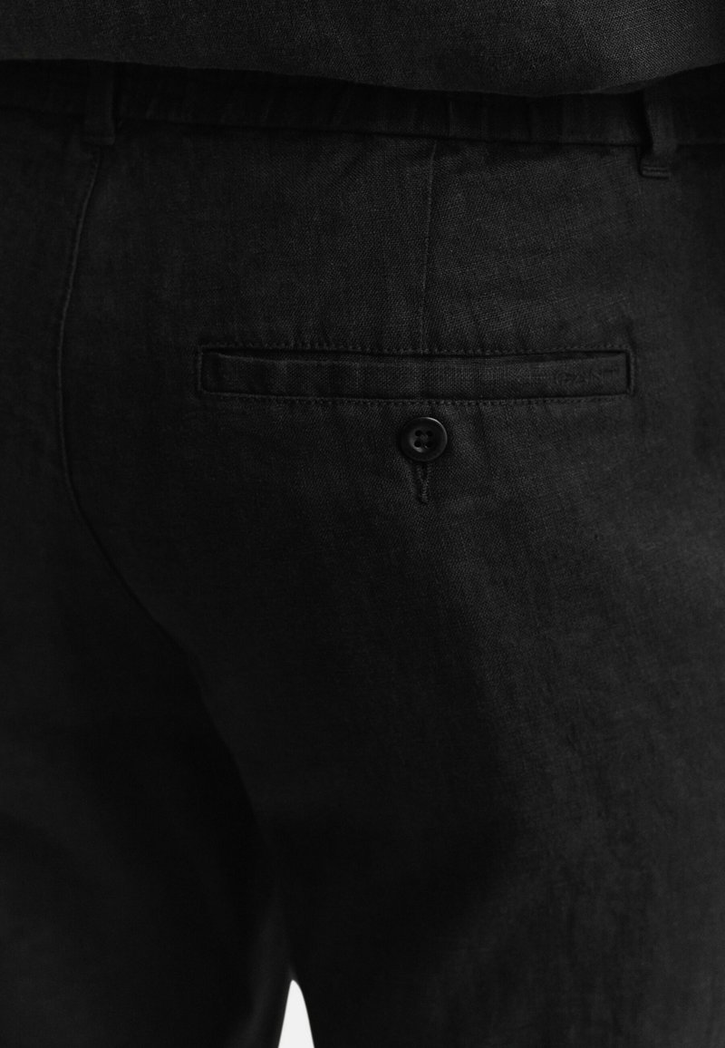 Gesäßtasche einer schwarzen Leinenhose mit einem einzelnen schwarzen Knopfverschluss, von der Seite an einer Person mit schwarzem Hemd gezeigt.
