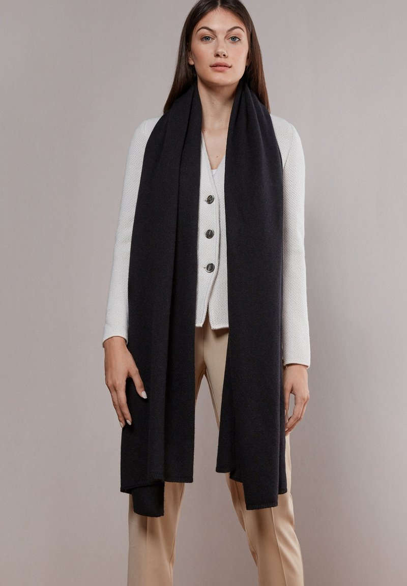 Sort uldhalstørklæde hænger ned fra skuldrene, parret med en knappet hvid sweater og neutrale bukser. Glat tekstur og elegant design.