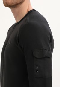 Zwarte lange mouwen shirt van gemetalliseerd stof, ronde halslijn en schouderpatch met twee metalen oogjes voor de afwerking.