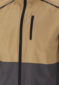 Leichte Jacke in Beige und Grau mit einem durchgehenden schwarzen Reißverschluss und einer Reißverschlusstasche auf der Brust. Glatte Textur und Farbschnitt-Design.
