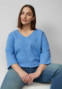 Blauwe, gestructureerde top met een V-hals en driekwart mouwen, gecombineerd met lichte denim jeans. Het materiaal heeft een subtiele geweven structuur.