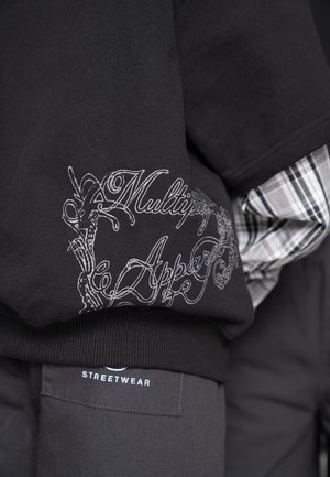 Sweat-shirt noir avec une inscription blanche ornée "Multipy Apper", superposé à une chemise à carreaux, accompagné d'un pantalon foncé étiqueté "Streetwear".