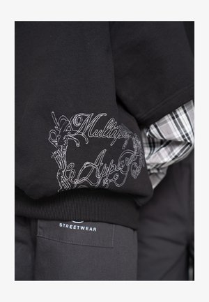 Sudadera negra con una elegante inscripción blanca que dice "Multipy Apper", combinada con una camisa de cuadros y pantalones oscuros etiquetados como "Streetwear".