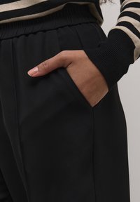 Pantalons noirs au toucher lisse, dotés de poches latérales et d'une taille élastique. Le bas de pantalon présente un détail côtelé dans une couleur contrastante.