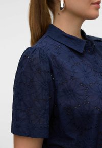 Camicia blu navy con colletto, maniche corte a sbuffo e ricami floreali. Presenta tagli e dettagli di superficie texture.