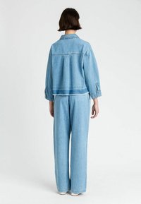 Giacca in denim azzurro chiaro con vestibilità rilassata, caratterizzata da un design con schiena plissettata e polsini con bottoni, abbinata a pantaloni in denim a gamba larga.