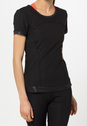 Žena v černém těsně přiléhajícím topu s krátkými rukávy a umělou koženou výztuží a černých legínách, zobrazený trup a levá ruka.