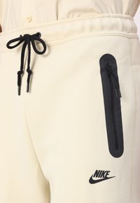 Κρέμα παντελόνια jogger με μαύρο κορδόνι και τσέπη με φερμουάρ, με το μαύρο λογότυπο της Nike στο μηρό. Μαλακό ύφασμα και άνετη εφαρμογή.