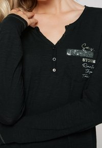 Zwarte lange mouw top met een gestructureerde afwerking, voorzien van een knoopsluiting bij de V-hals, een met pailletten versierde zak en geprinte tekstdetails.