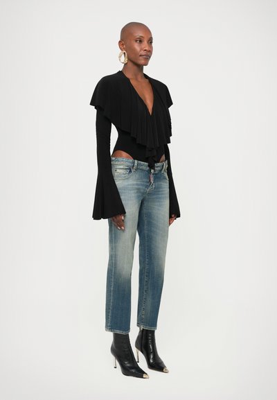 Γυναίκα με ξυρισμένο κεφάλι που φοράει μαύρο τοπ με βαθύ V και βολάν, ανοιχτόχρωμο μπλε τζιν, μαύρες μυτερές μπότες με ψηλό τακούνι και μεγάλα σκουλαρίκια, στέκεται πλευρικά.