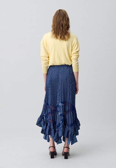 Gonna plissettata color navy con motivi floreali, orlo arricciato e pannelli semi-trasparenti. Abbinata a un maglione giallo. Sandali con tacco neri.