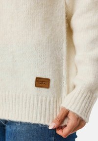 Kremvärd stickad tröja med ribbade ärmslut och nederkant. Har en brun läderetikett med "SWEDEMOUNT"-logotypen på framsidan. Mjuk textur.