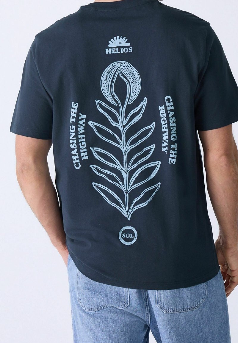 Vue de dos d'une personne portant un t-shirt noir avec un motif floral blanc et les textes "HELIOS," "CHASING THE HIGHWAY," et "SOL."