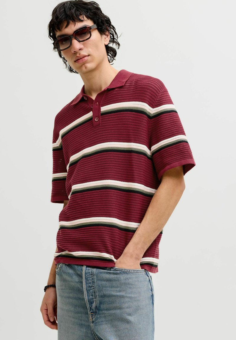 Jeune homme portant des lunettes de soleil, polo à manches courtes à rayures bordeaux, jean bleu clair, une main dans la poche.