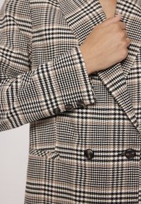 Dwurzędowa marynarka w beżowo-czarną białą kratę. Posiada szerokie klapy, teksturowany materiał i dwa czarne guziki na mankietach.