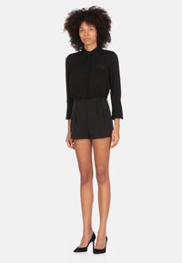 Blusa nera a maniche lunghe con colletto, vita cintrata, tasca frontale e pantaloni neri su misura, abbinati a tacchi alti neri.